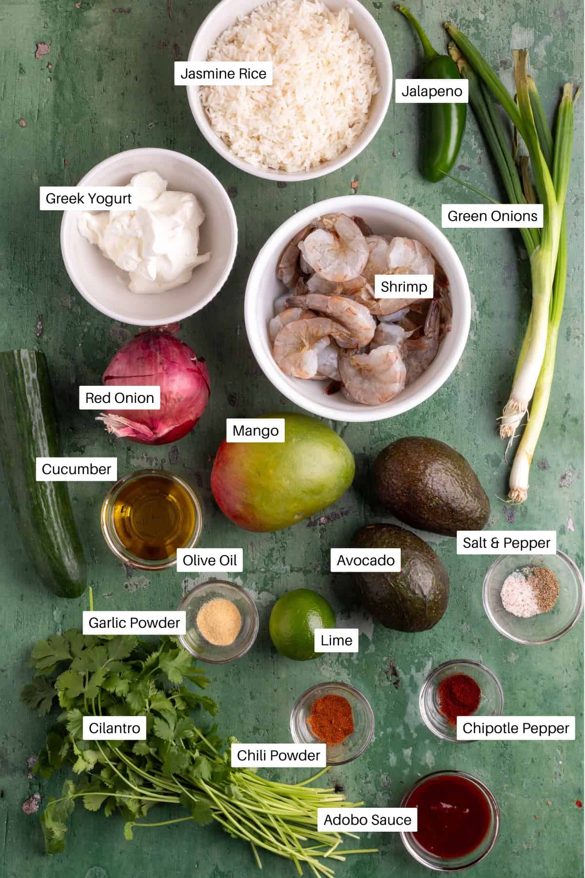 Top-down view of ingredients for a shrimp rice bowl on a green surface, featuring labeled bowls of jasmine rice, shrimp, Greek yogurt, red onion, green onions, jalapeño, cucumber, olive oil, mango, avocado, lime, spices, and herbs.