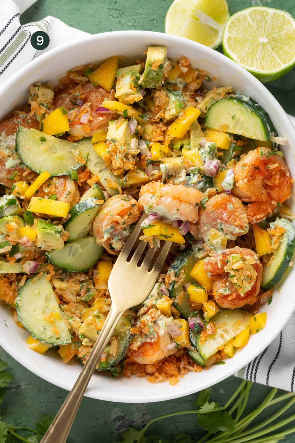 A vibrant shrimp rice bowl features sliced cucumbers, diced mango, avocado, red onion, and rice, garnished with herbs. A gold fork rests on the salad while lime halves sit nearby on a fresh green surface.