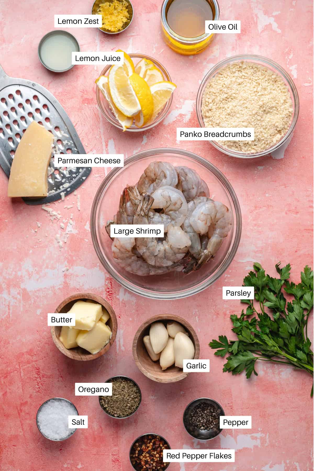 A top view of ingredients for shrimp oreganata on a pink surface, including large shrimp, lemon zest, lemon juice, olive oil, panko breadcrumbs, Parmesan cheese, parsley, butter, garlic, oregano, salt, pepper, and red pepper flakes.