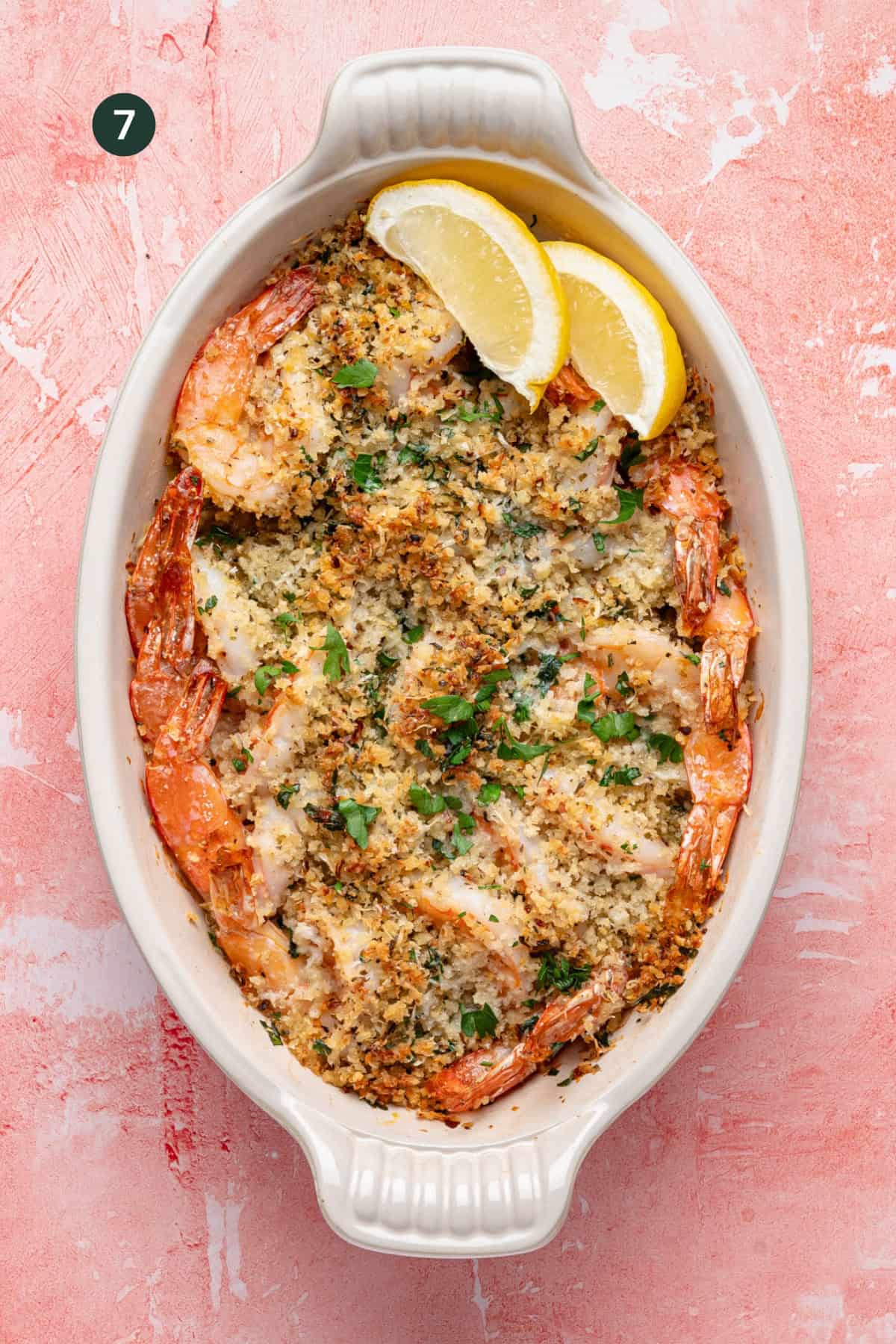 An oval baking dish filled with shrimp oreganata, topped with a golden breadcrumb crust, garnished with chopped parsley, and served with two lemon wedges on a pink textured background.