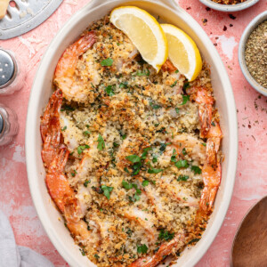An oval baking dish filled with shrimp oreganata, topped with golden breadcrumbs and herbs, garnished with two lemon wedges. Small bowls of red pepper flakes and spices surround the dish on a pink surface.