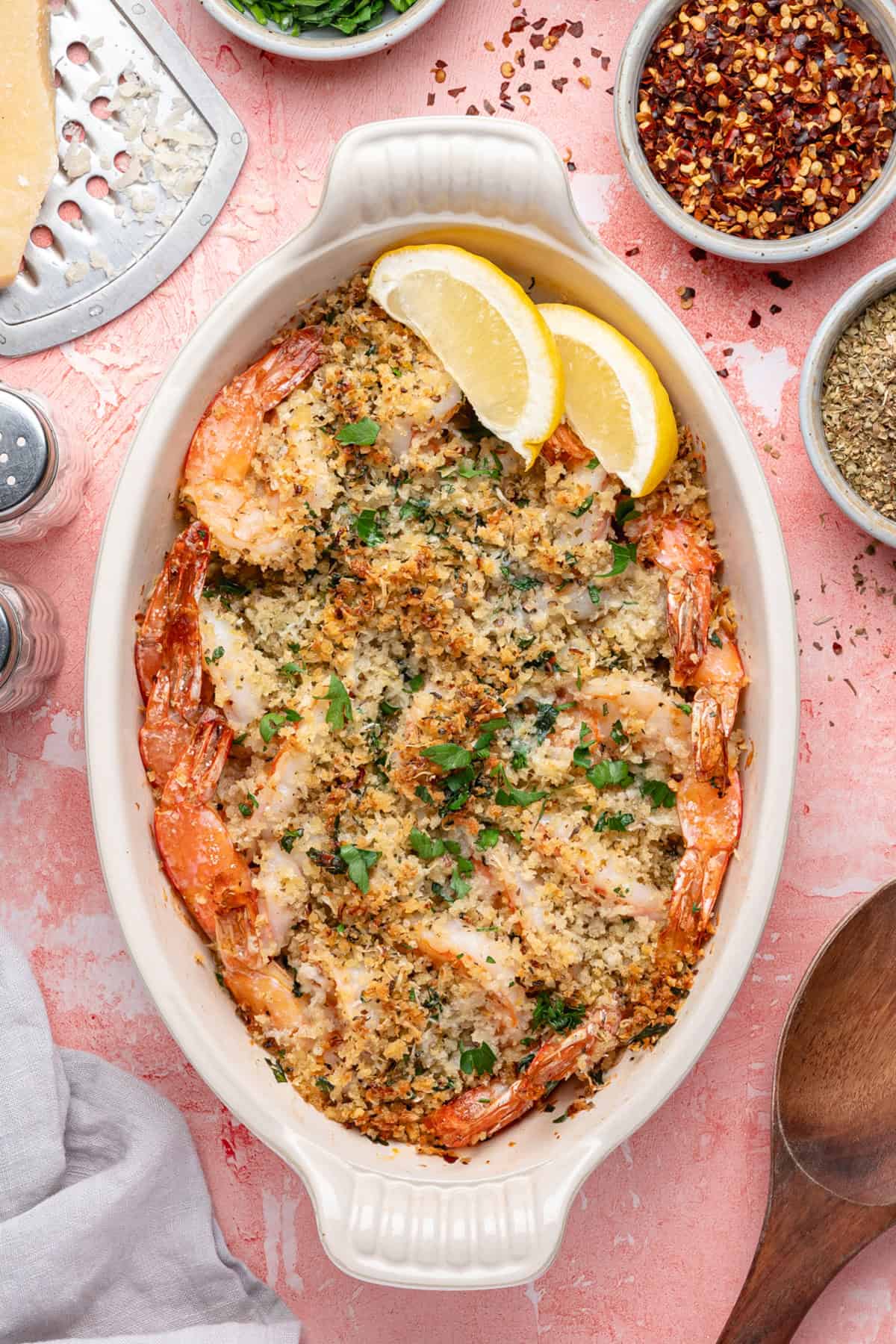An oval baking dish filled with shrimp oreganata, topped with golden breadcrumbs and herbs, garnished with two lemon wedges. Small bowls of red pepper flakes and spices surround the dish on a pink surface.