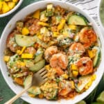 A white bowl filled with a vibrant shrimp rice bowl—shrimp, sliced avocado, cucumber, mango, red onion, herbs, and creamy dressing—sits on a green surface with lime wedges and fresh cilantro. A striped napkin and fork are beside the bowl.