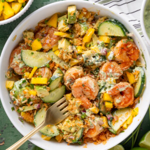 A white bowl filled with a vibrant shrimp rice bowl—shrimp, sliced avocado, cucumber, mango, red onion, herbs, and creamy dressing—sits on a green surface with lime wedges and fresh cilantro. A striped napkin and fork are beside the bowl.
