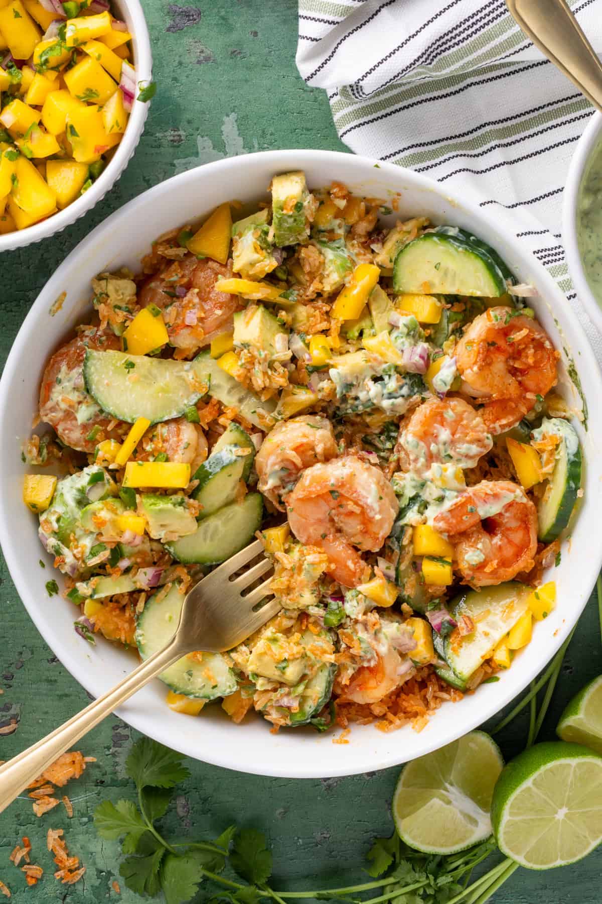 A white bowl filled with a vibrant shrimp rice bowl—shrimp, sliced avocado, cucumber, mango, red onion, herbs, and creamy dressing—sits on a green surface with lime wedges and fresh cilantro. A striped napkin and fork are beside the bowl.