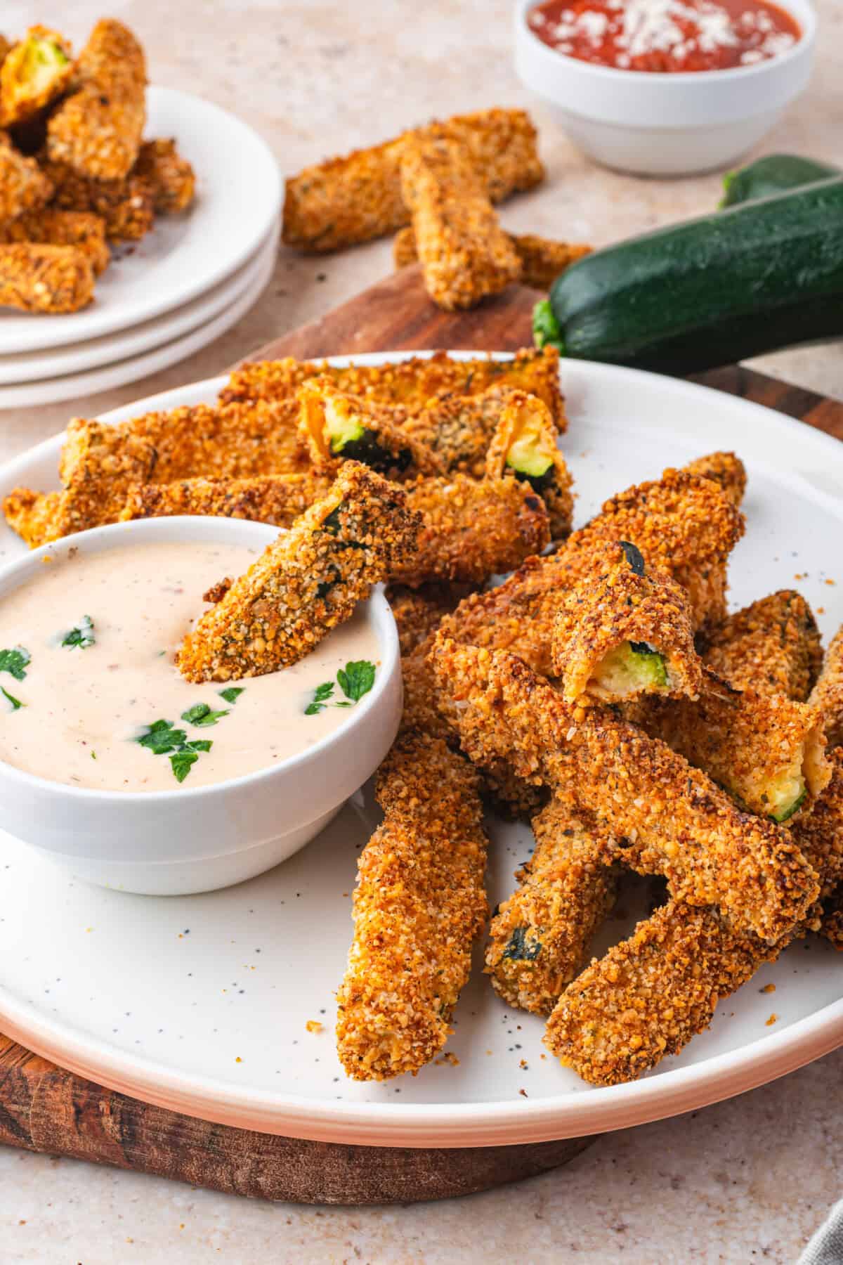 A plate of crispy air fryer zucchini fries is served with a bowl of creamy dipping sauce, garnished with herbs. More fries and a zucchini are in the background, alongside a bowl of red sauce.