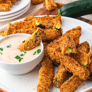 A plate of crispy air fryer zucchini fries is served with a bowl of creamy dipping sauce, garnished with herbs. More fries and a zucchini are in the background, alongside a bowl of red sauce.