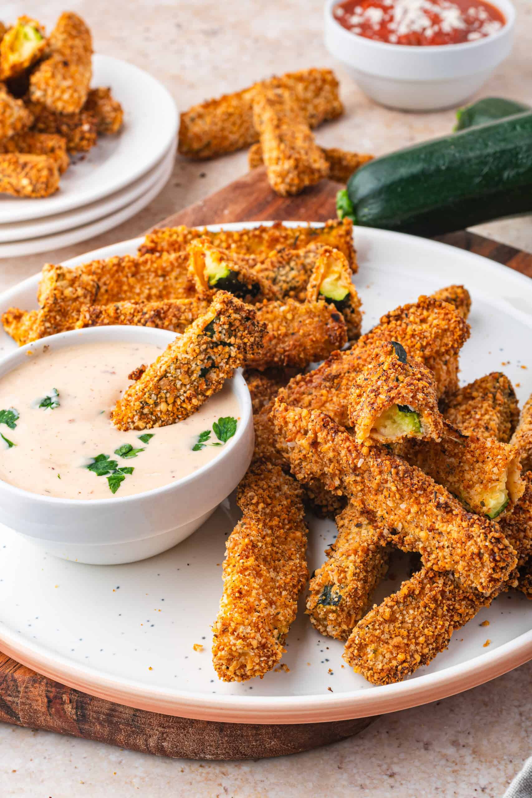A plate of crispy air fryer zucchini fries is served with a bowl of creamy dipping sauce, garnished with herbs. More fries and a zucchini are in the background, alongside a bowl of red sauce.