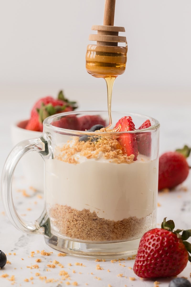 A glass mug filled with No Bake Cheesecake Cups, layered and topped with strawberries, blueberries, and crumbs, as honey drizzles from a dipper above. Whole strawberries and crumbs are scattered around.