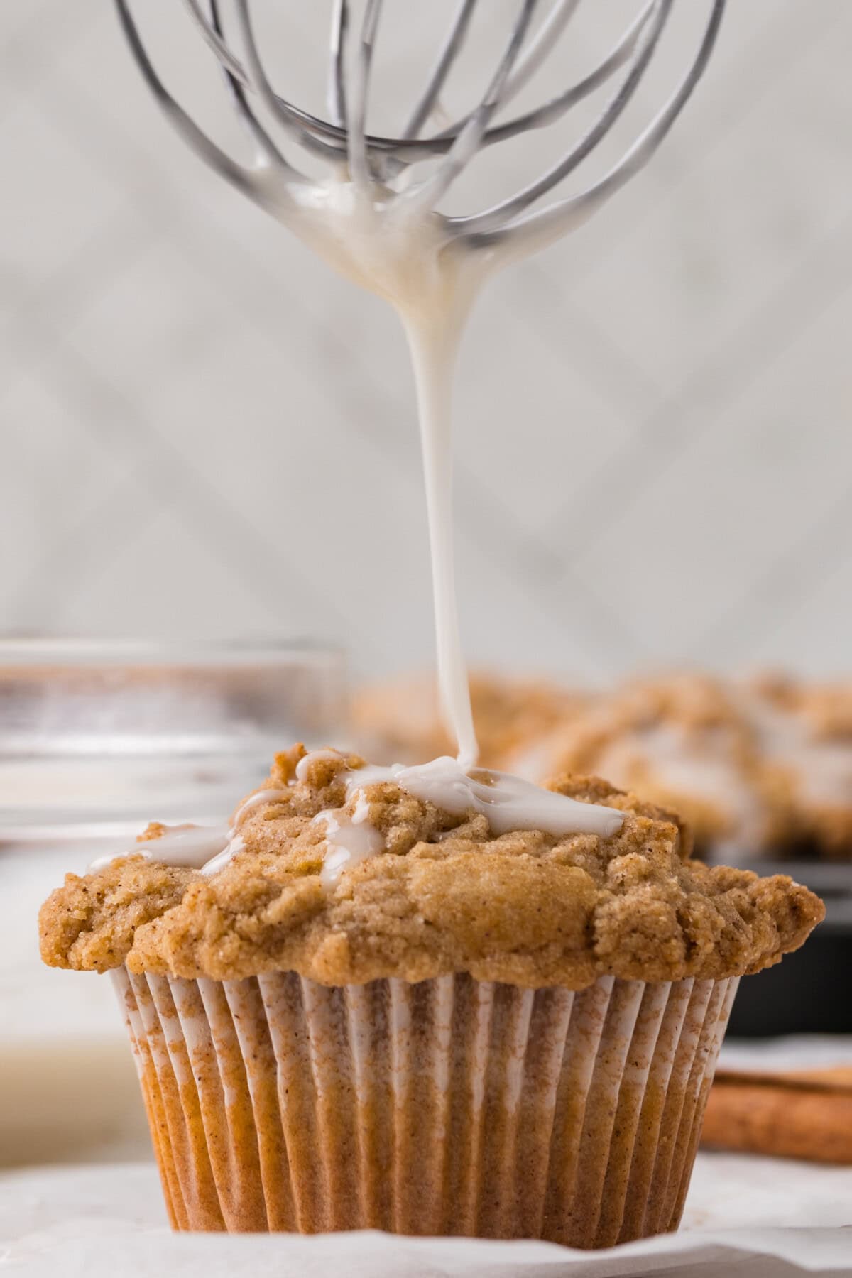 A close-up of a Coffee Cake Muffin with a crumbly topping, as white icing is drizzled onto it from a whisk above. In the background, another Coffee Cake Muffin and blurred kitchen items are visible.