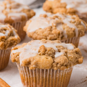 Four Coffee Cake Muffins with crumbly cinnamon streusel tops and a light glaze are arranged on a white surface, with a cinnamon stick in the foreground and a striped cloth in the background.