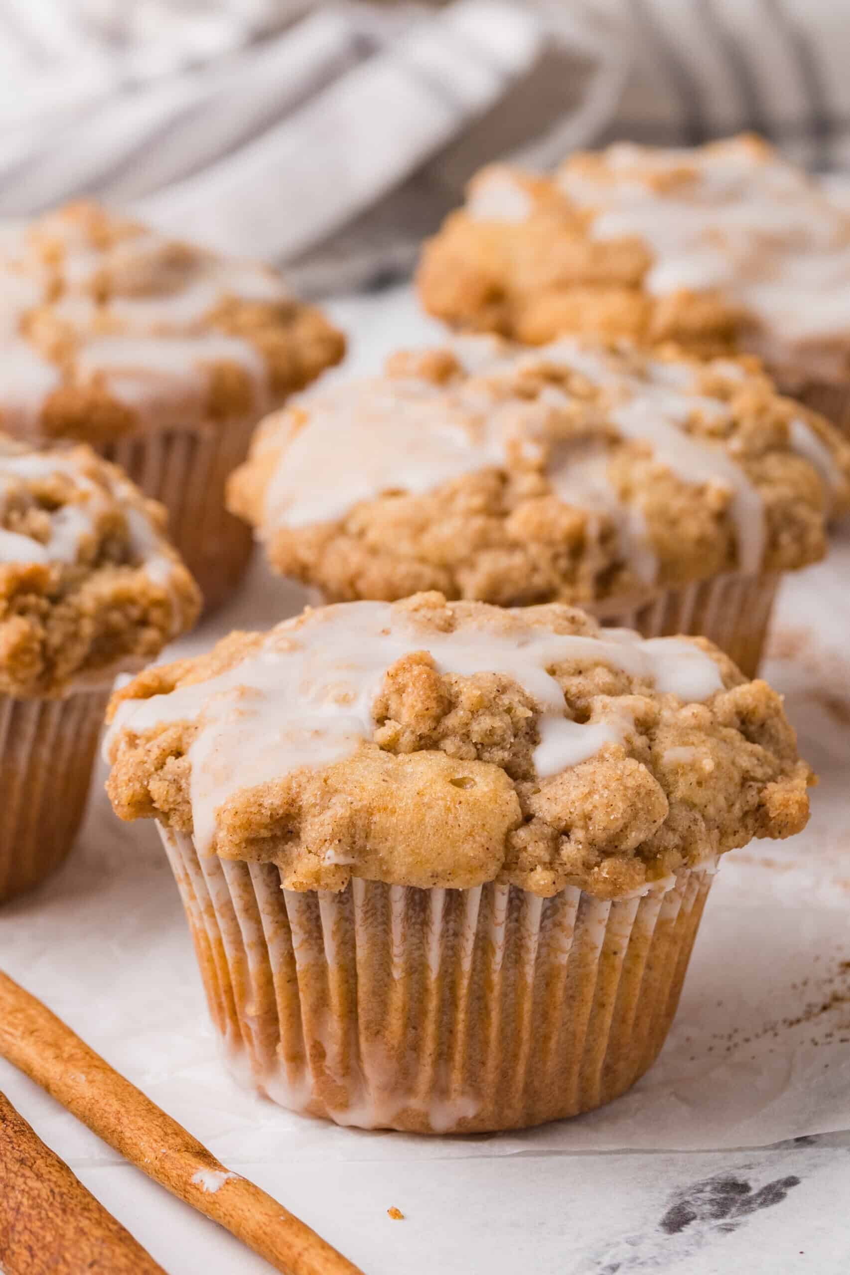 Four Coffee Cake Muffins with crumbly cinnamon streusel tops and a light glaze are arranged on a white surface, with a cinnamon stick in the foreground and a striped cloth in the background.