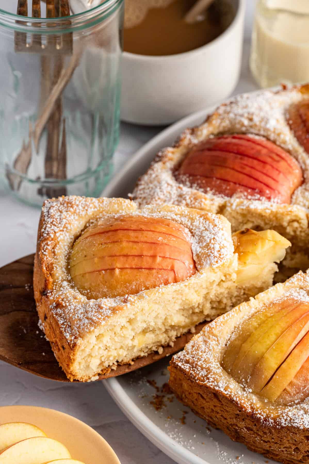 A close-up of a sliced German Apple Cake Recipe with powdered sugar on top. A wooden spatula lifts one slice, revealing a moist interior. Glasses and a cup of coffee are in the background.