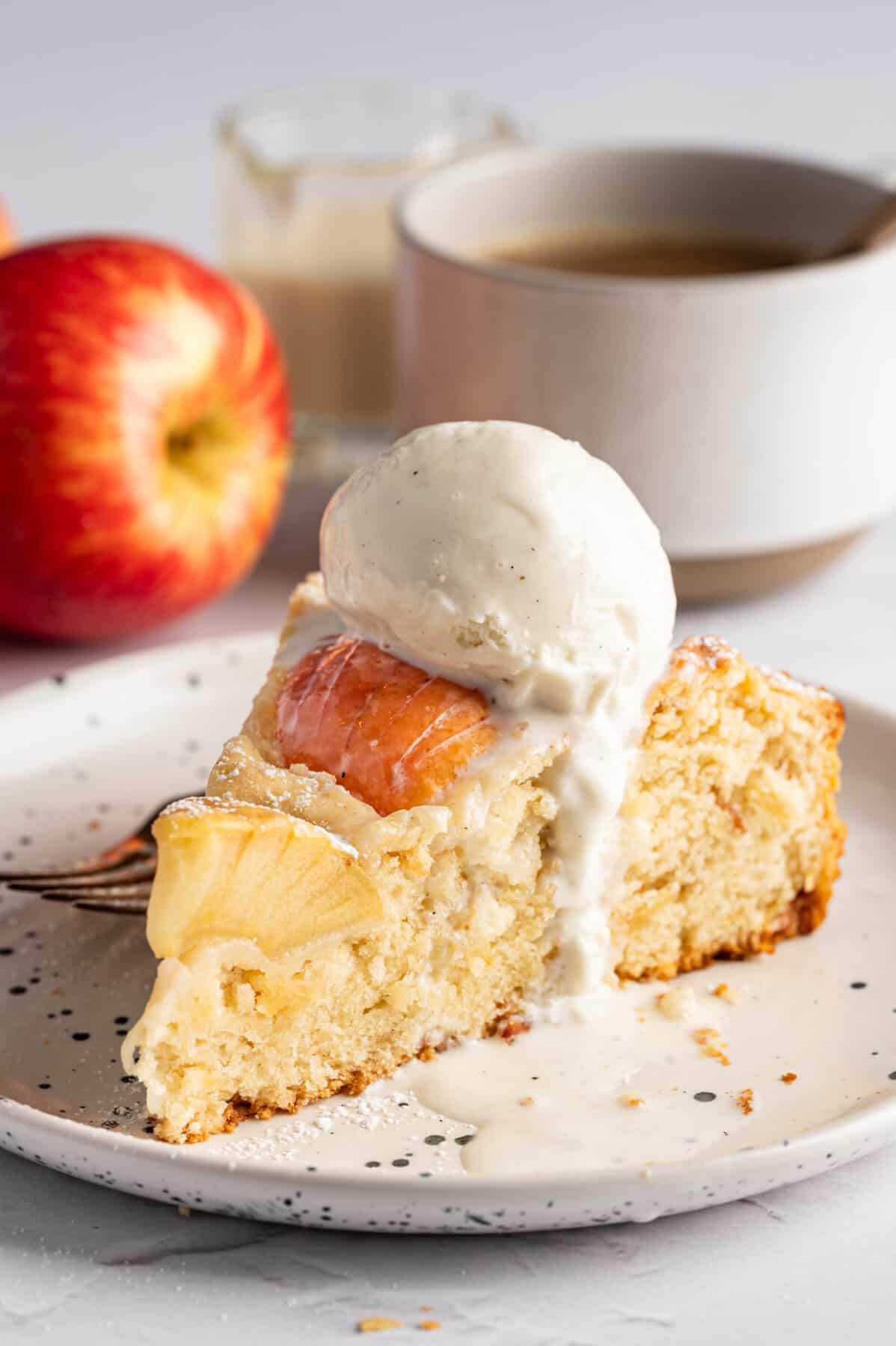 A slice of German Apple Cake topped with vanilla ice cream rests on a plate beside a fork. In the background are a fresh apple, a cup of coffee, and a small glass of cream.