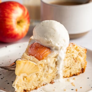 A slice of German Apple Cake topped with vanilla ice cream rests on a plate beside a fork. In the background are a fresh apple, a cup of coffee, and a small glass of cream.