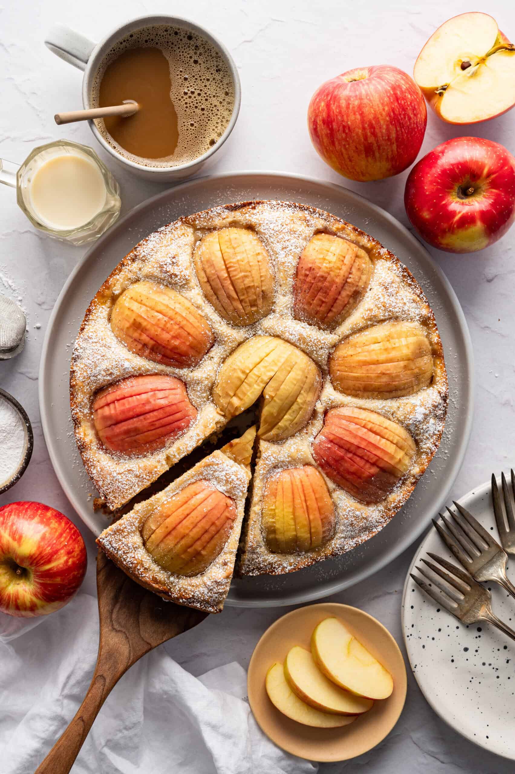 A round German Apple Cake dusted with powdered sugar sits on a plate, one slice being served. Fresh apples, coffee, milk, apple slices, and plates with forks surround this tasty German Apple Cake recipe.