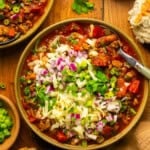 A bowl of pork chili topped with shredded cheese, chopped red onion, and green onions sits on a wooden table, surrounded by fresh bread, extra toppings, and cilantro.