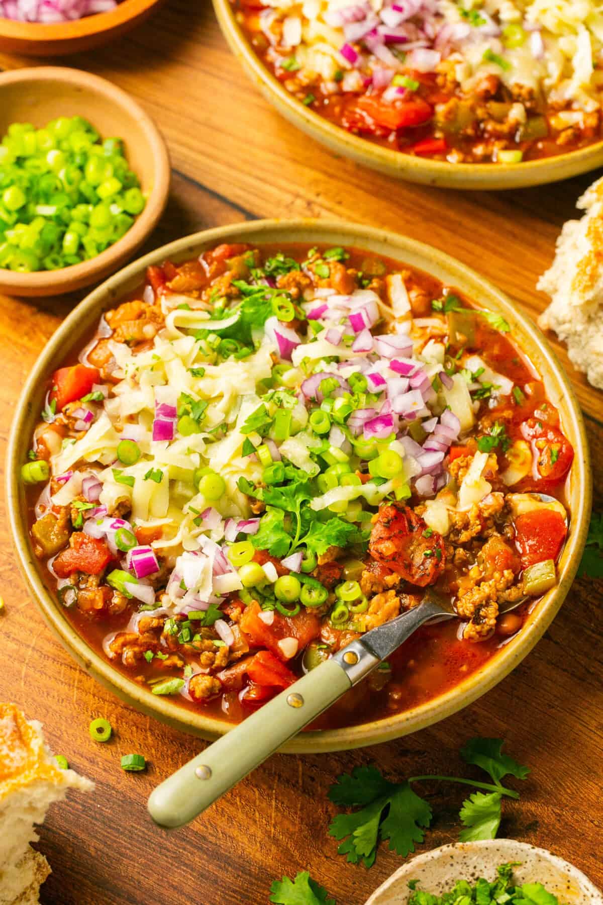 A bowl of pork chili topped with chopped red onions, shredded cheese, green onions, and cilantro, with a spoon inside. Surrounding the bowl are extra green onions, bread, and fresh cilantro on a wooden table.