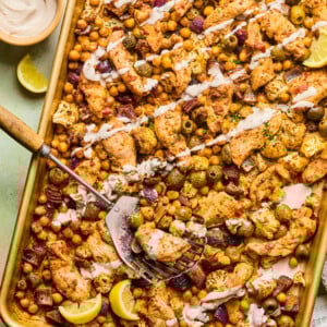 A harissa tray bake chicken sheet pan filled with roasted chickpeas, cauliflower, olives, and red onion, drizzled with a creamy sauce and garnished with lemon wedges. A spatula rests on the pan, ready to serve.