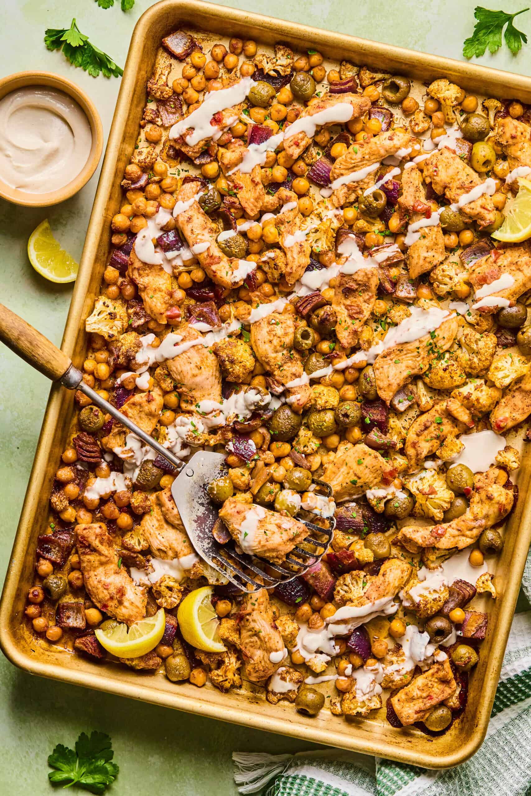 A harissa tray bake chicken sheet pan filled with roasted chickpeas, cauliflower, olives, and red onion, drizzled with a creamy sauce and garnished with lemon wedges. A spatula rests on the pan, ready to serve.