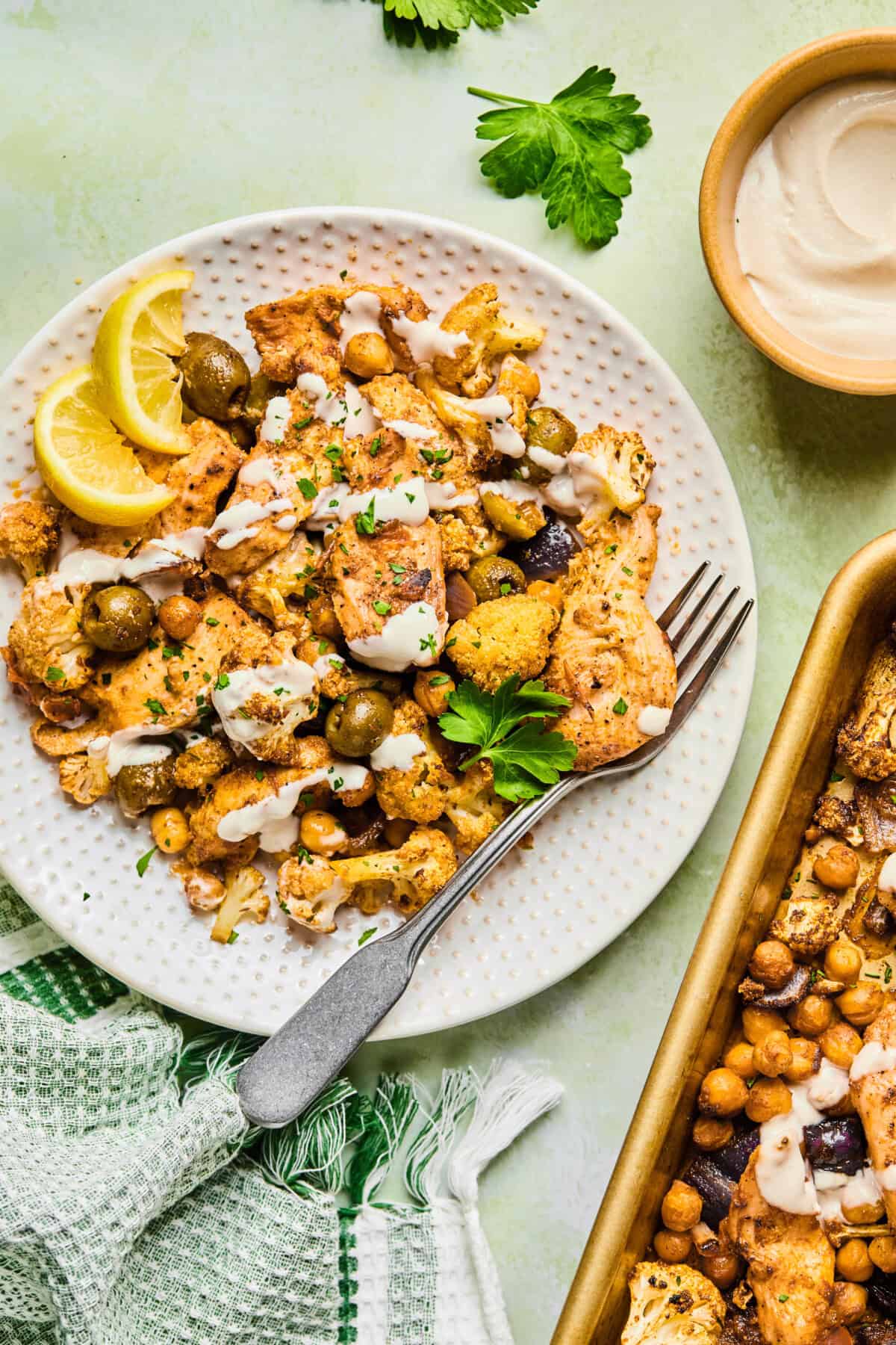 A plate of roasted cauliflower, chickpeas, olives, and tofu—reminiscent of a vibrant harissa tray bake chicken—is garnished with parsley and lemon wedges beside a baking tray and creamy sauce. A fork and napkin rest nearby on the green surface.