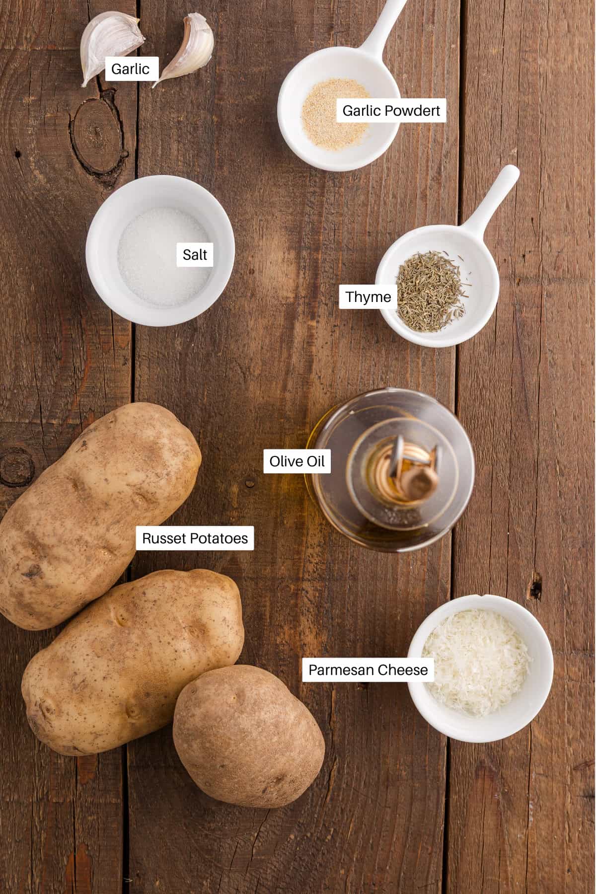 Overhead view of ingredients for potato wedges in the air fryer: russet potatoes, grated parmesan cheese, olive oil, thyme, garlic powder, salt, and garlic cloves arranged on a wooden surface with each item labeled.