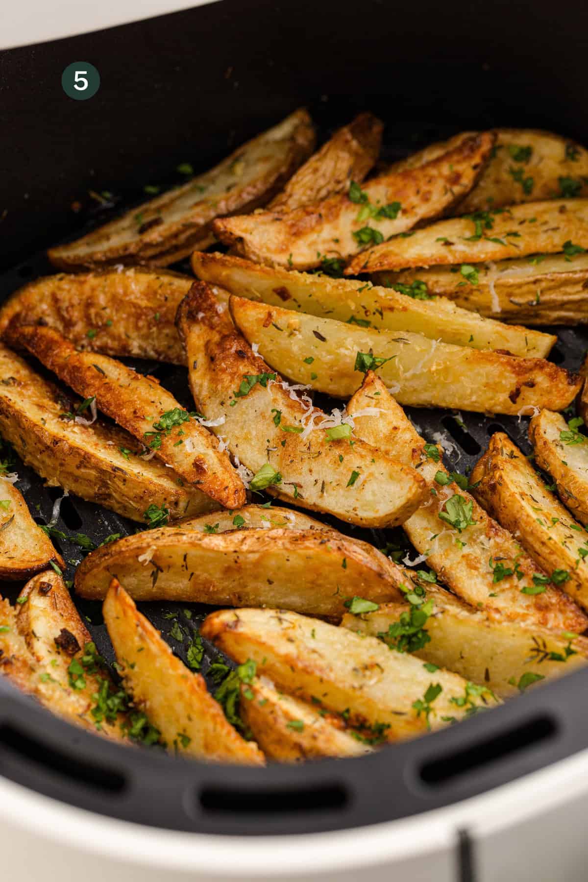 Golden potato wedges in the air fryer, garnished with herbs and grated cheese, are arranged in a single layer inside the basket.