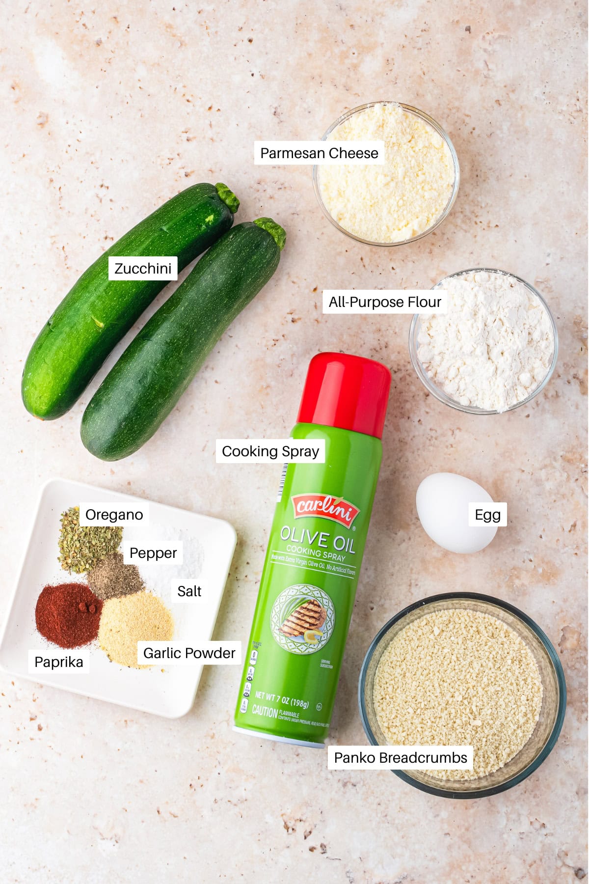 Top view of ingredients for air fryer zucchini fries, including two zucchinis, parmesan cheese, all-purpose flour, panko breadcrumbs, an egg, cooking spray, and small bowls of oregano, pepper, salt, paprika, and garlic powder.