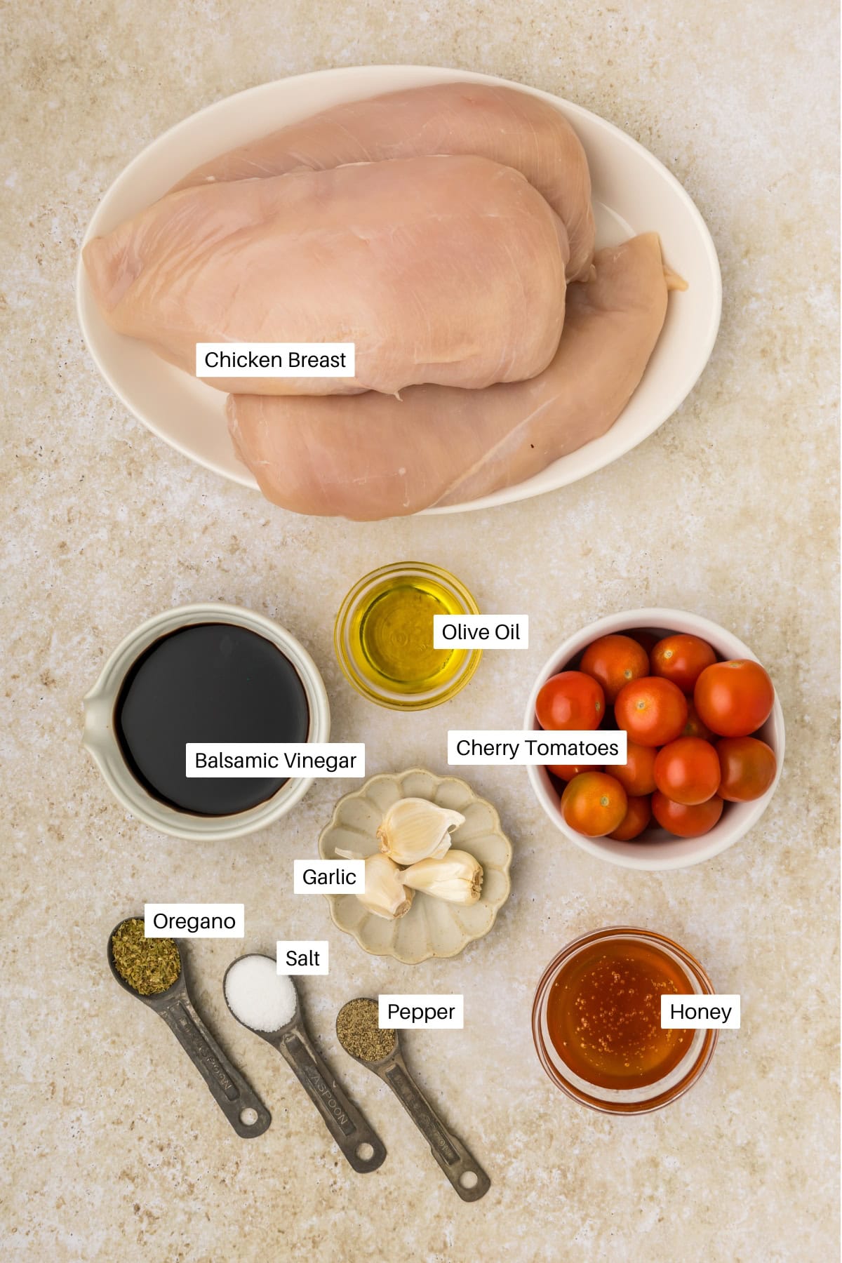 Two raw chicken breasts on a plate, surrounded by labeled bowls of cherry tomatoes, olive oil, balsamic vinegar, honey, garlic cloves, and measuring spoons of oregano, salt, and pepperโperfect for an easy balsamic chicken recipe.
