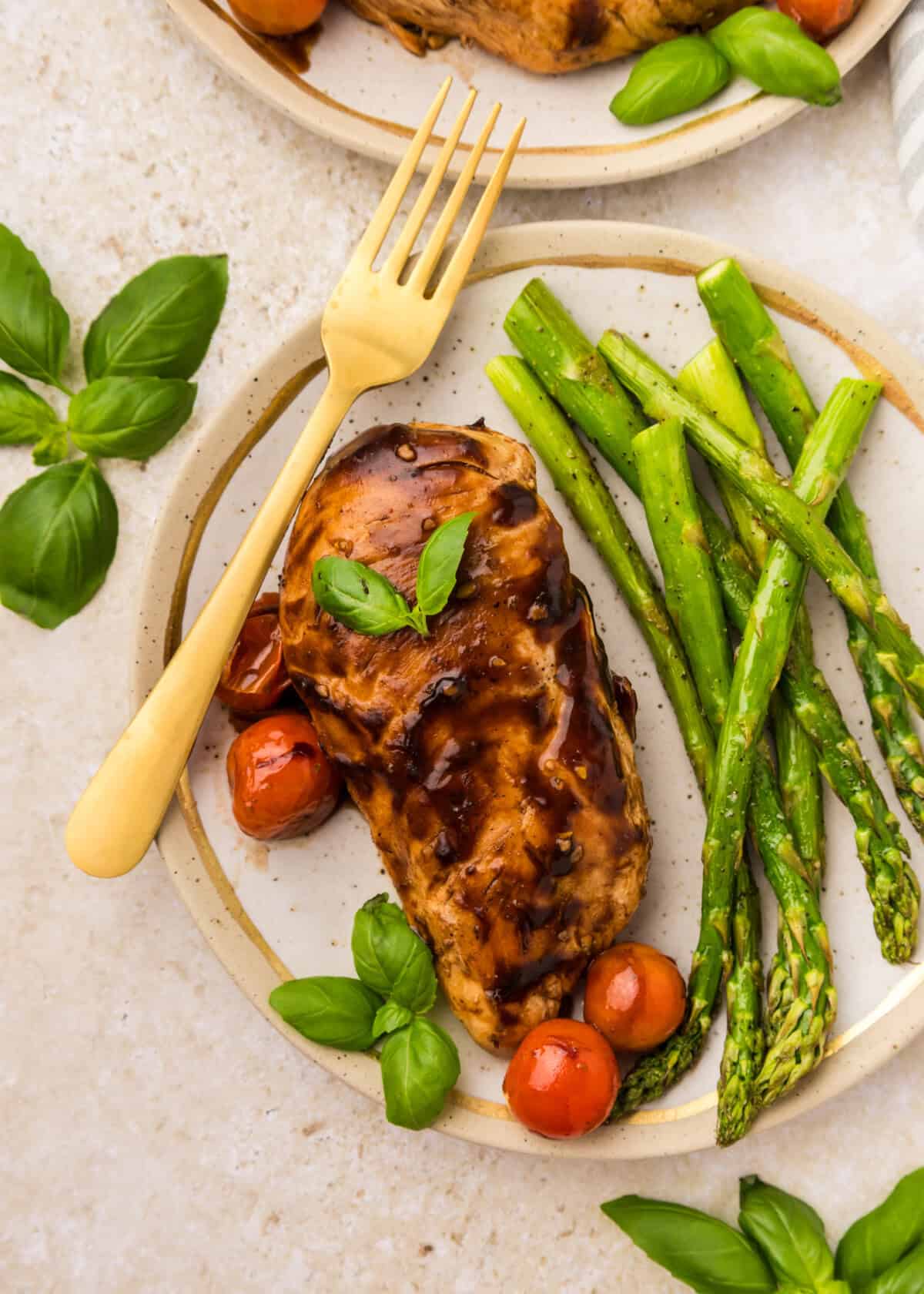 A plate with balsamic-glazed chicken breast, roasted asparagus spears, and cherry tomatoesโpart of a delicious balsamic chicken recipeโgarnished with fresh basil leaves. A gold fork rests on the plate.