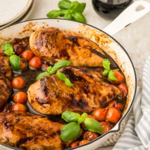 Boneless chicken breasts cooked in a skillet with cherry tomatoes and fresh basil leaves, drizzled with a dark balsamic glaze. This balsamic chicken recipe is served with a bottle of balsamic vinegar, plate, and napkin nearby.