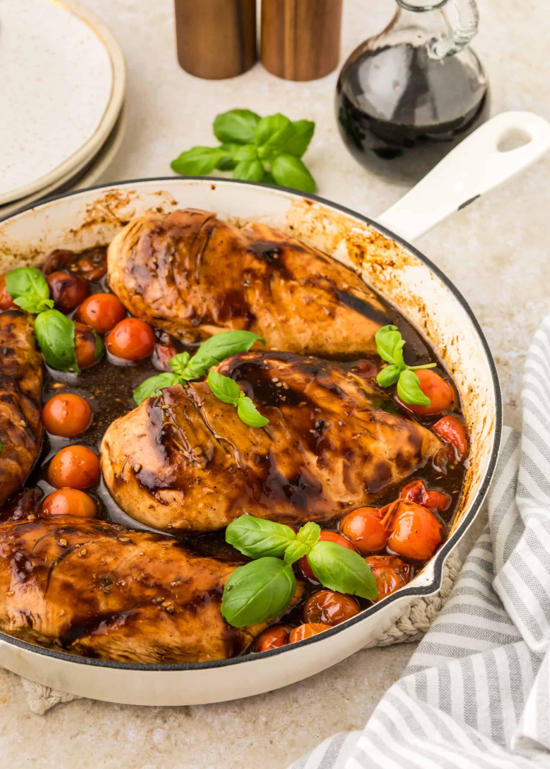 Boneless chicken breasts cooked in a skillet with cherry tomatoes and fresh basil leaves, drizzled with a dark balsamic glaze. This balsamic chicken recipe is served with a bottle of balsamic vinegar, plate, and napkin nearby.