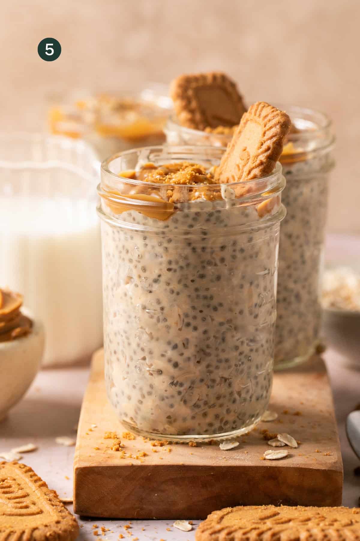 Two glass jars filled with creamy biscoff overnight oats, topped with crushed cookies and a cookie piece. The jars rest on a wooden board, surrounded by a glass of milk and extra cookies in the background.