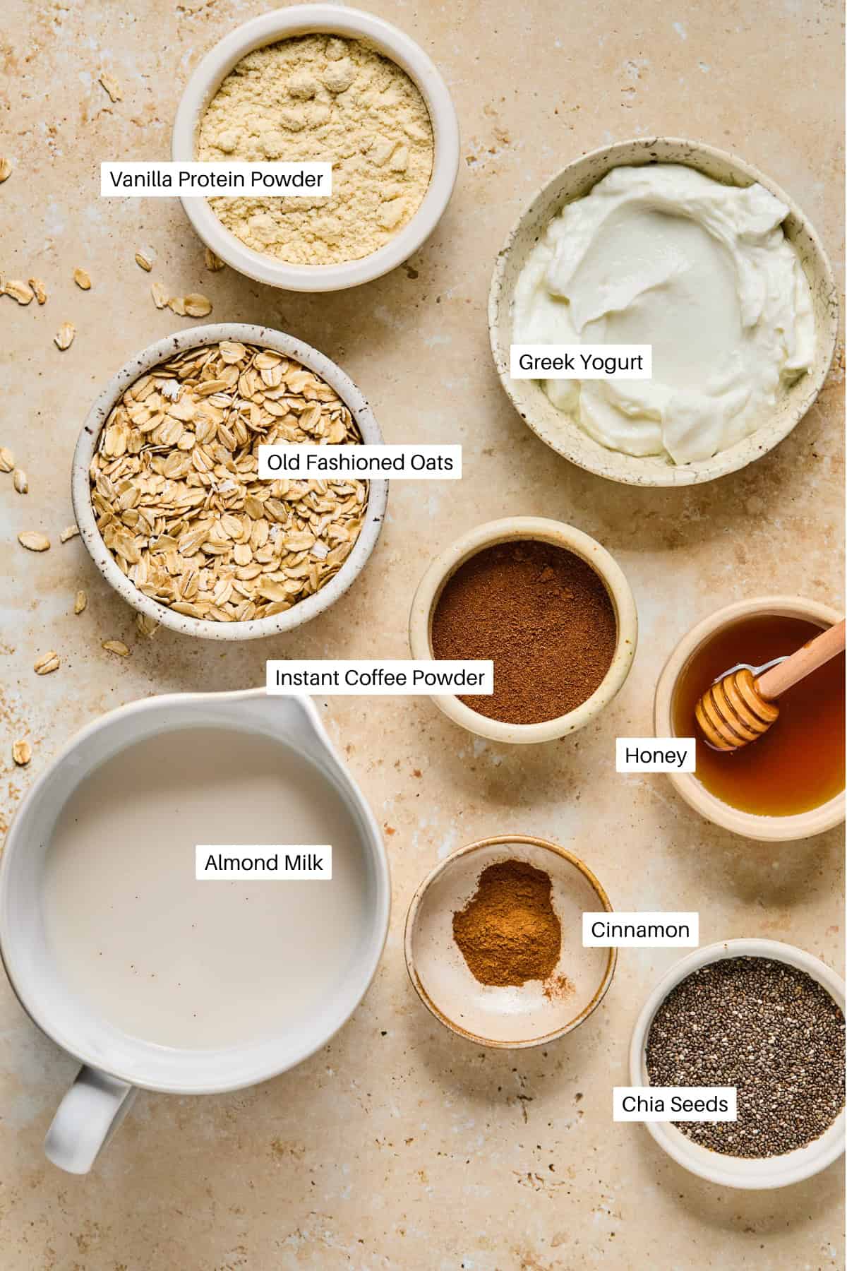 A top-down view of bowls containing labeled ingredients for coffee overnight oats: vanilla protein powder, Greek yogurt, old fashioned oats, almond milk, instant coffee powder, honey, cinnamon, and chia seeds on a beige surface.