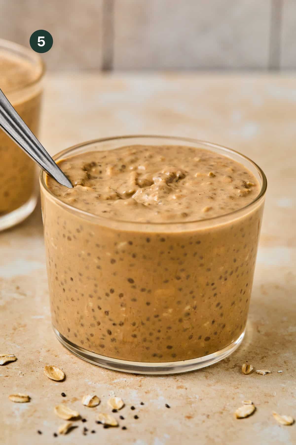 A glass jar filled with creamy, light brown coffee overnight oats mixed with chia seeds, with a metal spoon inside. Some oats are scattered on the tan surface nearby. Another jar is partially visible in the background.