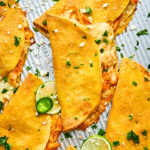 Golden, crispy chicken tacos filled with shredded chicken and melted cheese, garnished with fresh cilantro, sliced jalapeños, lime wedges, and crumbled cheese on a textured metal tray.