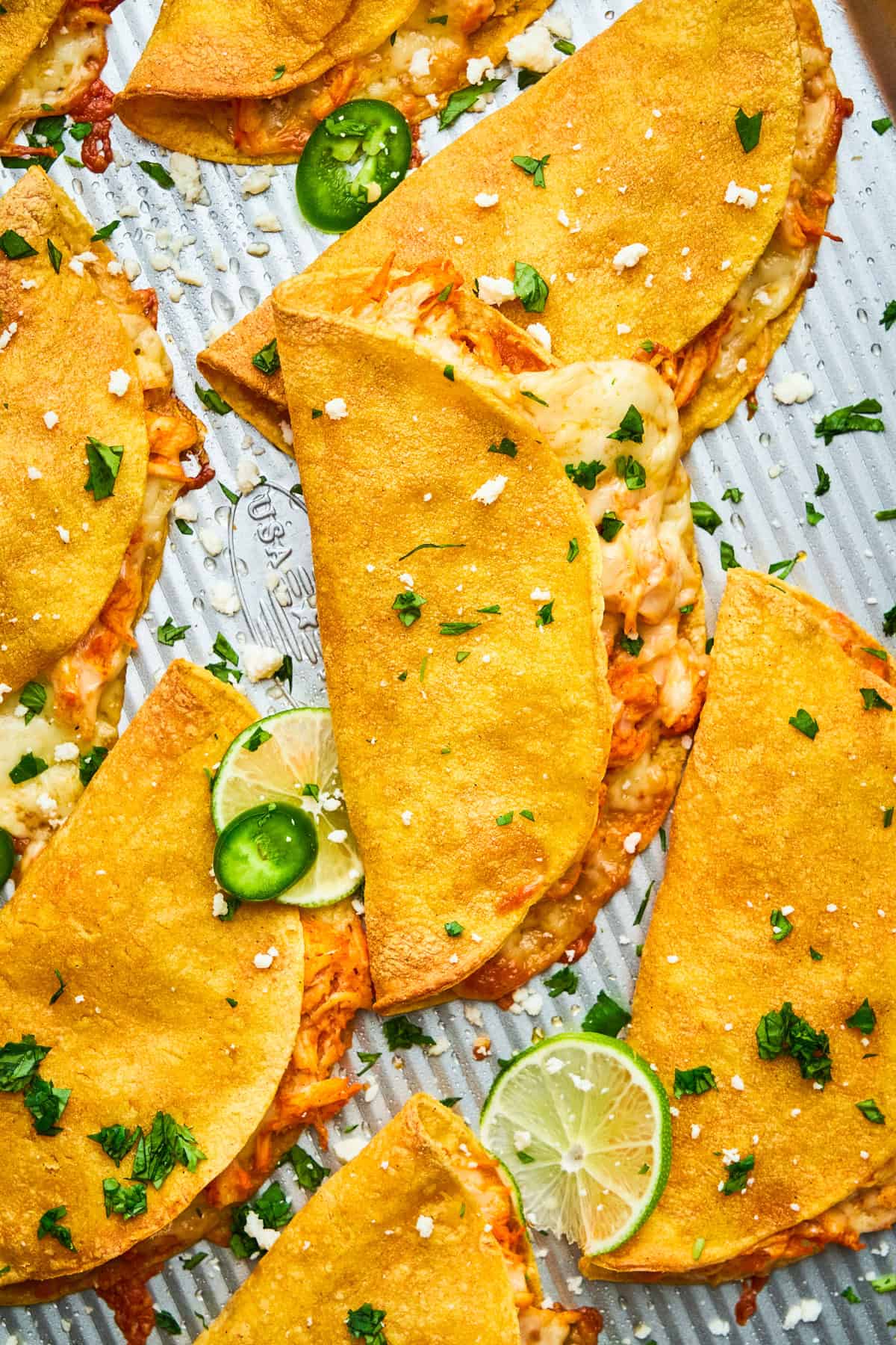Golden, crispy chicken tacos filled with shredded chicken and melted cheese, garnished with fresh cilantro, sliced jalapeños, lime wedges, and crumbled cheese on a textured metal tray.