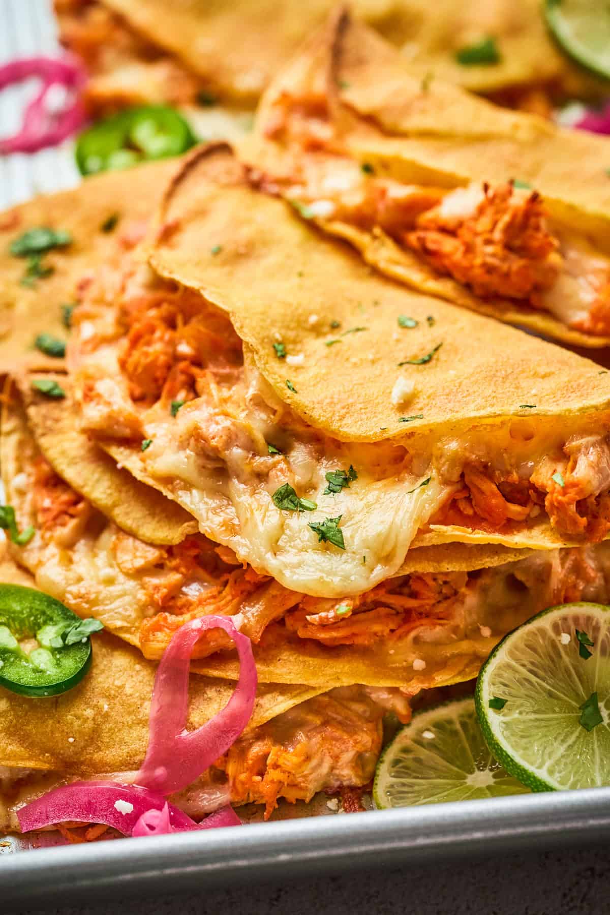 Close-up of crispy quesadillas filled with shredded chicken and melted cheese, reminiscent of crispy chicken tacos, garnished with chopped cilantro. Slices of jalapeño, pickled onions, and lime wedges are visible on the side.