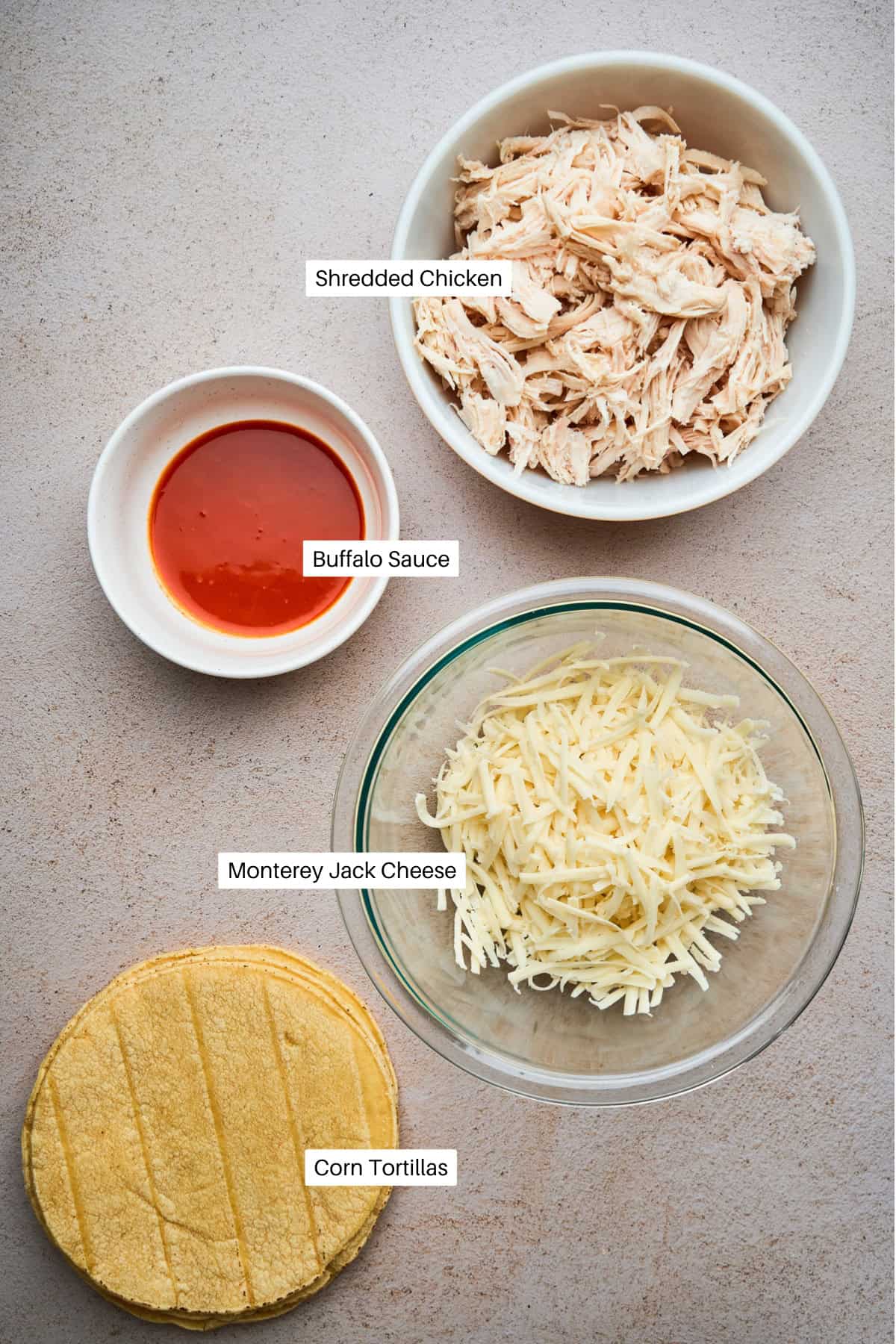 Four bowls and plates of ingredients for crispy chicken tacos on a light surface: shredded chicken, buffalo sauce, shredded Monterey Jack cheese, and a stack of corn tortillas, each labeled with text.