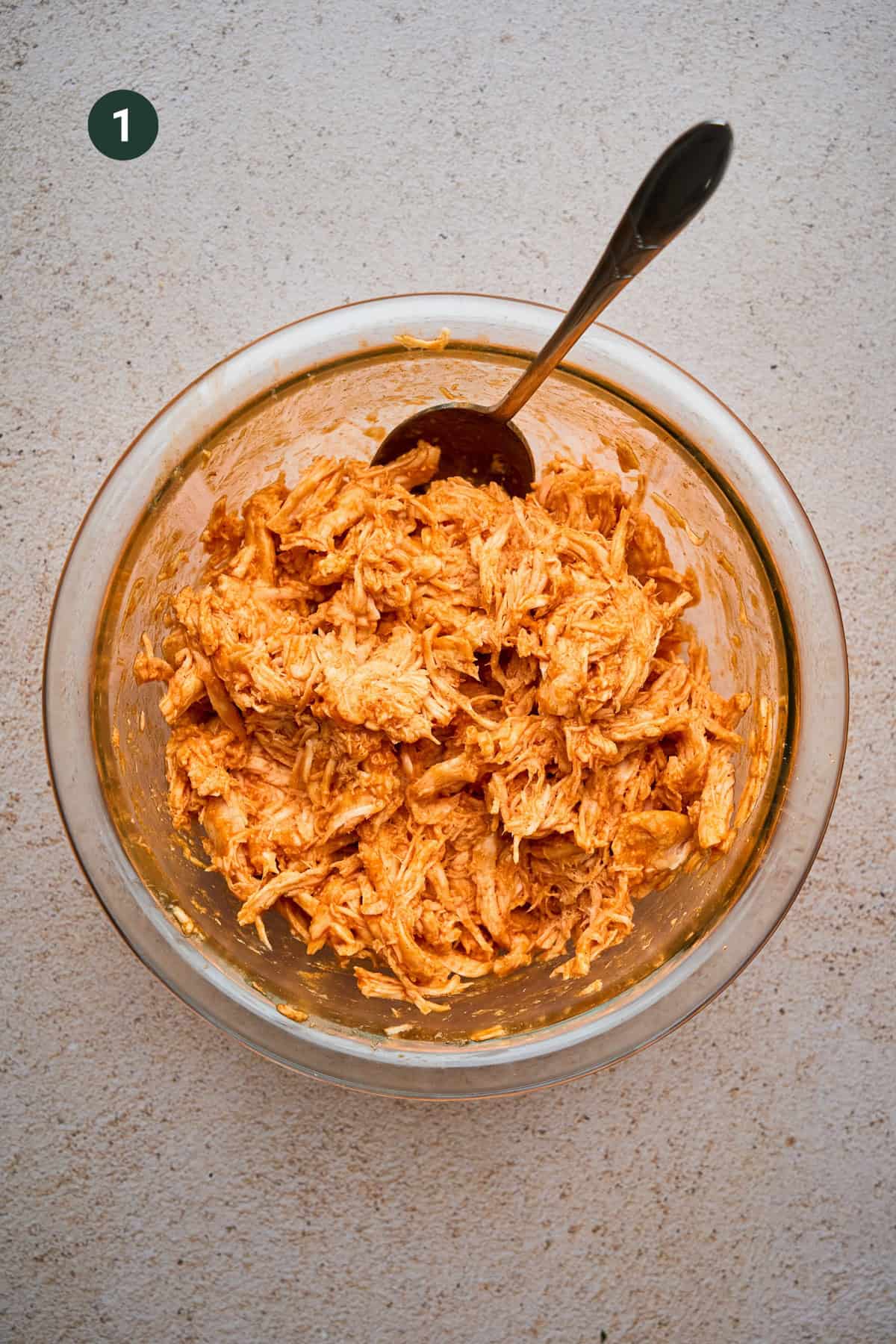 A glass bowl filled with shredded chicken mixed with an orange sauce, perfect for crispy chicken tacos, sits on a light-colored surface. A spoon rests in the bowl, and a green circle with the number 1 is in the top left corner.