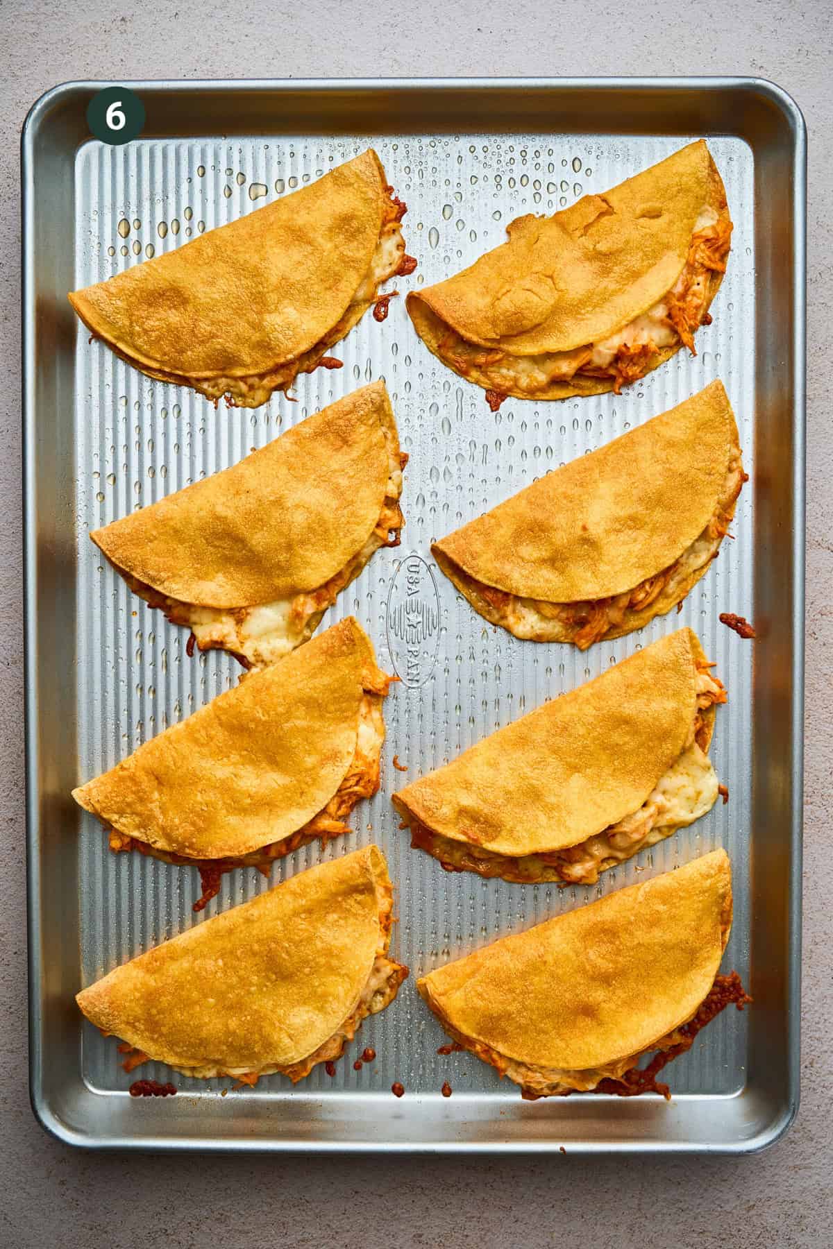 A baking tray with eight folded, golden-brown crispy chicken tacos filled with melted cheese and shredded chicken, arranged in neat rows on a light countertop.