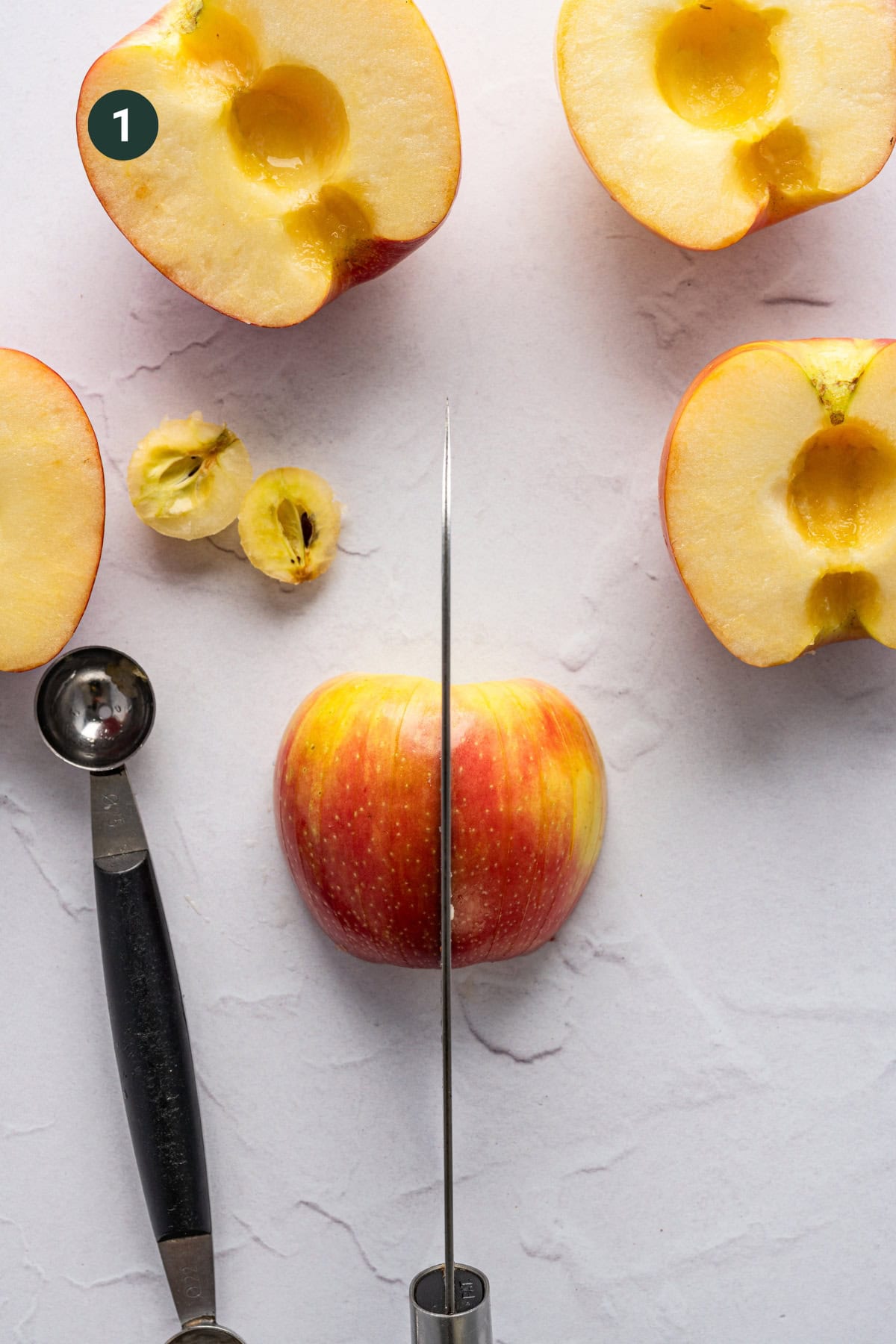 A fresh apple cut into halves and quarters is arranged on a white surface, ready for a German Apple Cake Recipe. A metal melon baller and a knife stand beside the apple, with two seeds and apple cores displayed nearby.
