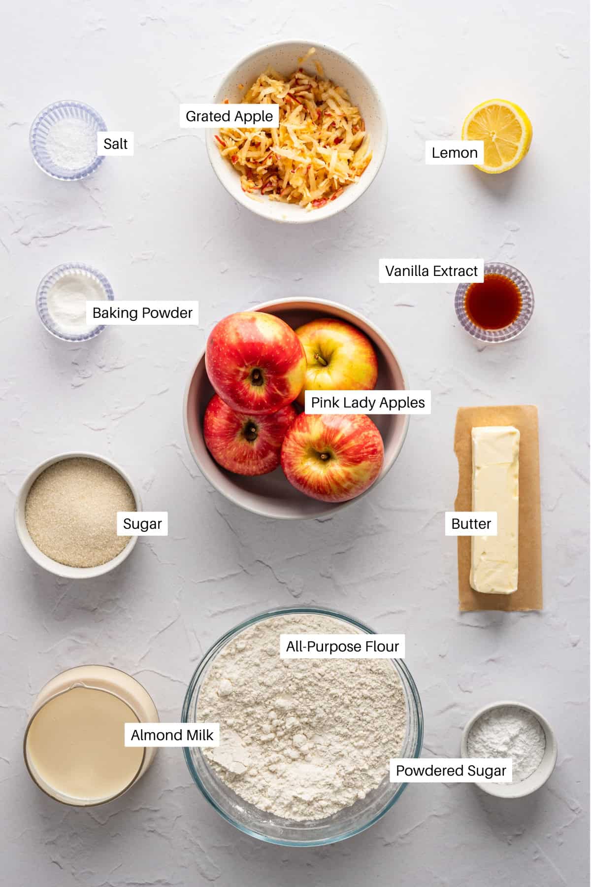 Overhead view of baking ingredients for a German Apple Cake Recipe on a white surface, including Pink Lady apples, grated apple, lemon half, butter, vanilla extract, almond milk, flour, sugars, baking powder, and saltโeach item clearly labeled.