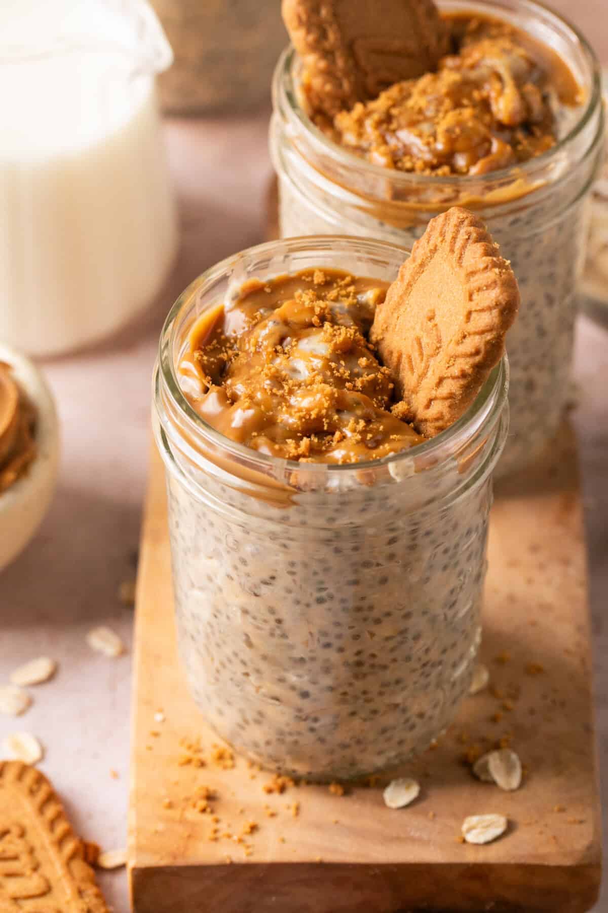 Two glass jars filled with chia pudding topped with caramel sauce, crumbled cookie pieces, and a whole cookie sit on a wooden boardโperfect alongside a cup of milk and scattered oats for fans of biscoff overnight oats.