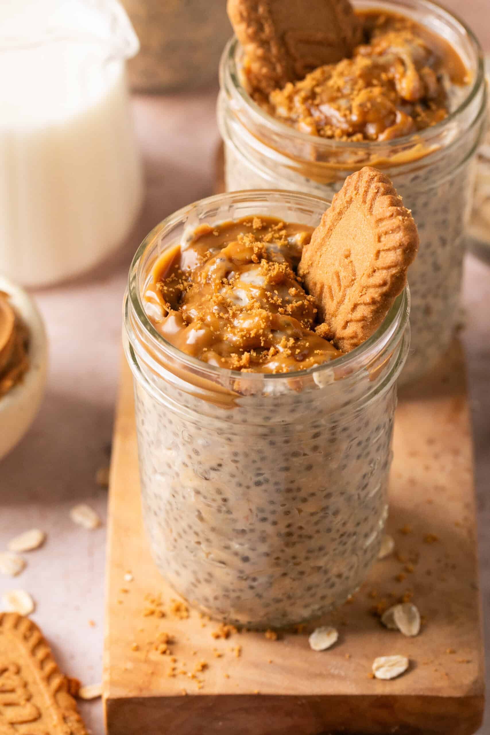Two glass jars filled with chia pudding topped with caramel sauce, crumbled cookie pieces, and a whole cookie sit on a wooden board—perfect alongside a cup of milk and scattered oats for fans of biscoff overnight oats.