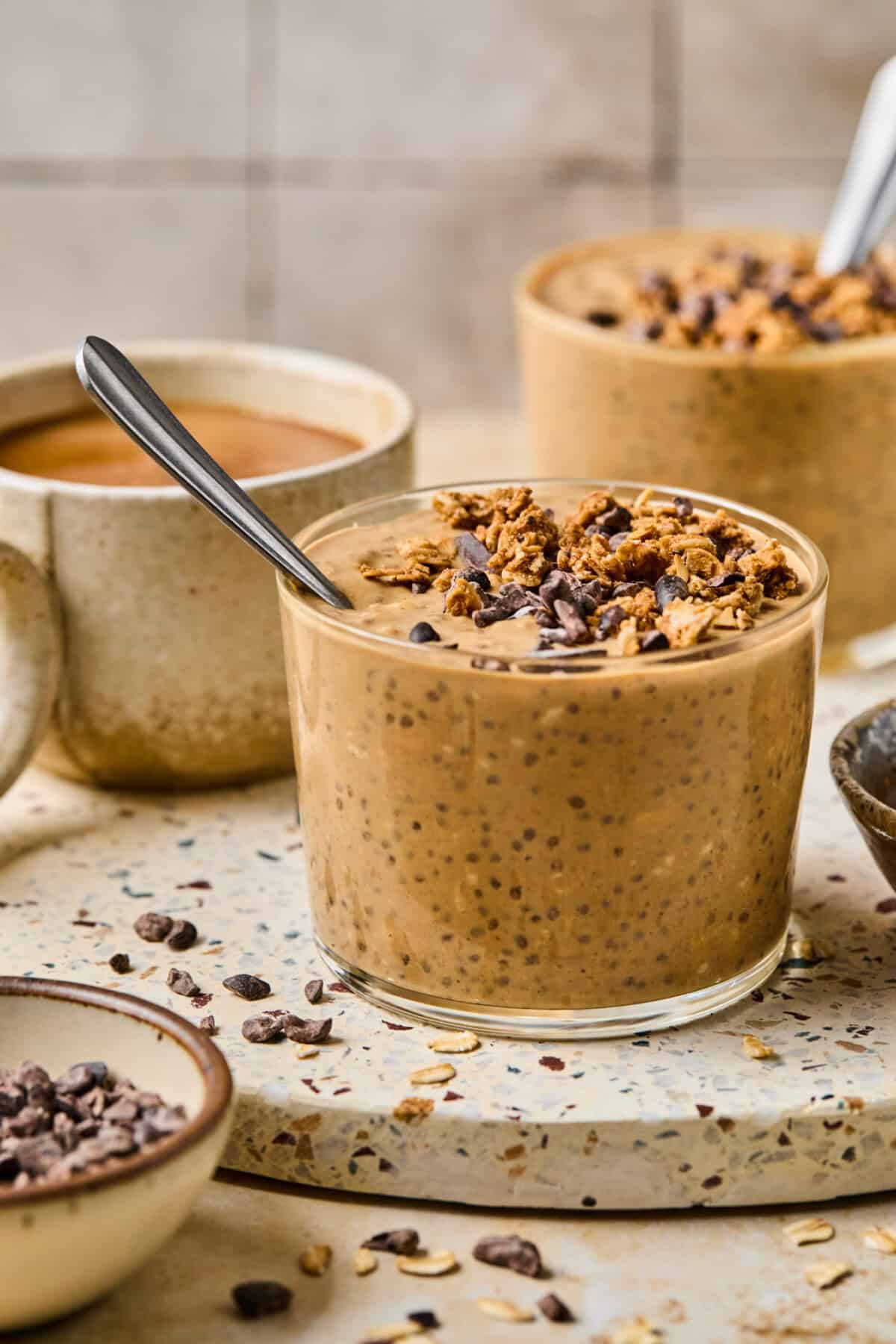 A glass filled with creamy chia pudding, topped with granola and cacao nibs, sits on a speckled surface. Spoons are in the glasses, while mugs and bowls of coffee overnight oats and similar ingredients are nearby.