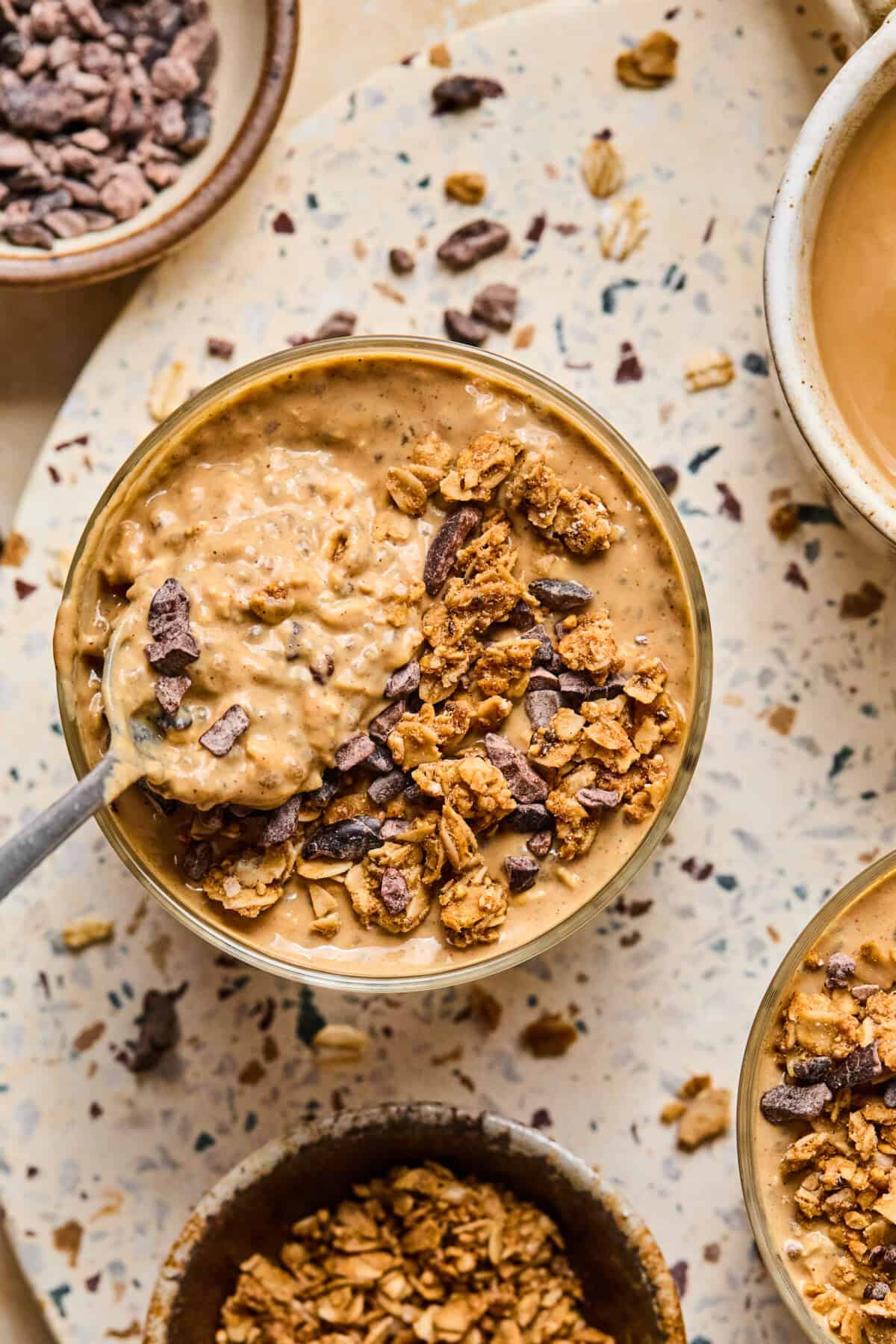A bowl of creamy coffee overnight oats topped with granola and chocolate chunks sits on a speckled surface, surrounded by smaller bowls of granola and a beverage. A spoon rests in the oatmeal.