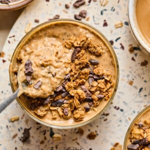 A bowl of creamy coffee overnight oats topped with granola and chocolate chunks sits on a speckled surface, surrounded by smaller bowls of granola and a beverage. A spoon rests in the oatmeal.