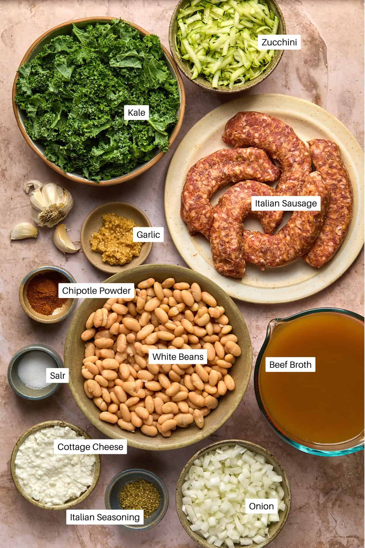 Bowls with labeled ingredients for Sausage and White Bean Soup, including kale, shredded zucchini, Italian sausage, garlic, chipotle powder, salt, white beans, beef broth, cottage cheese, Italian seasoning, and chopped onion.