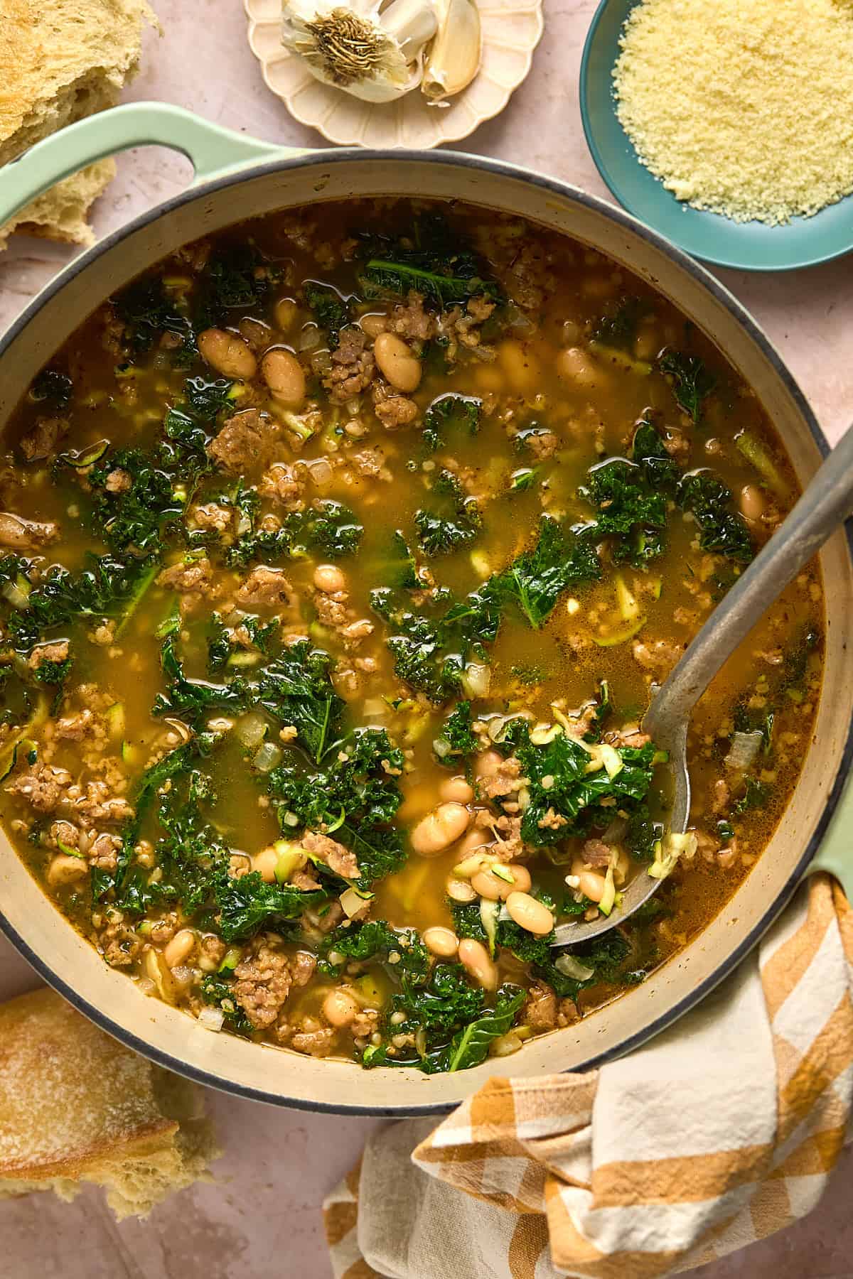 A large pot of Sausage and White Bean Soup with kale simmers in broth. A ladle rests inside the pot. Nearby are a plate of grated cheese, a garlic clove, and pieces of crusty bread on a beige table.